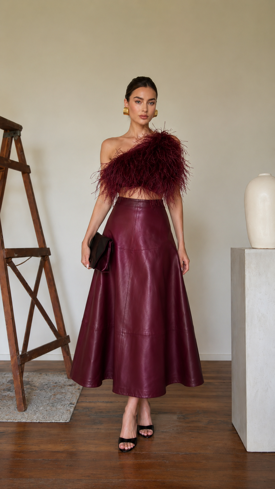Model wearing a burgundy high-waisted faux leather A-line midi skirt with a one-shoulder feather top
